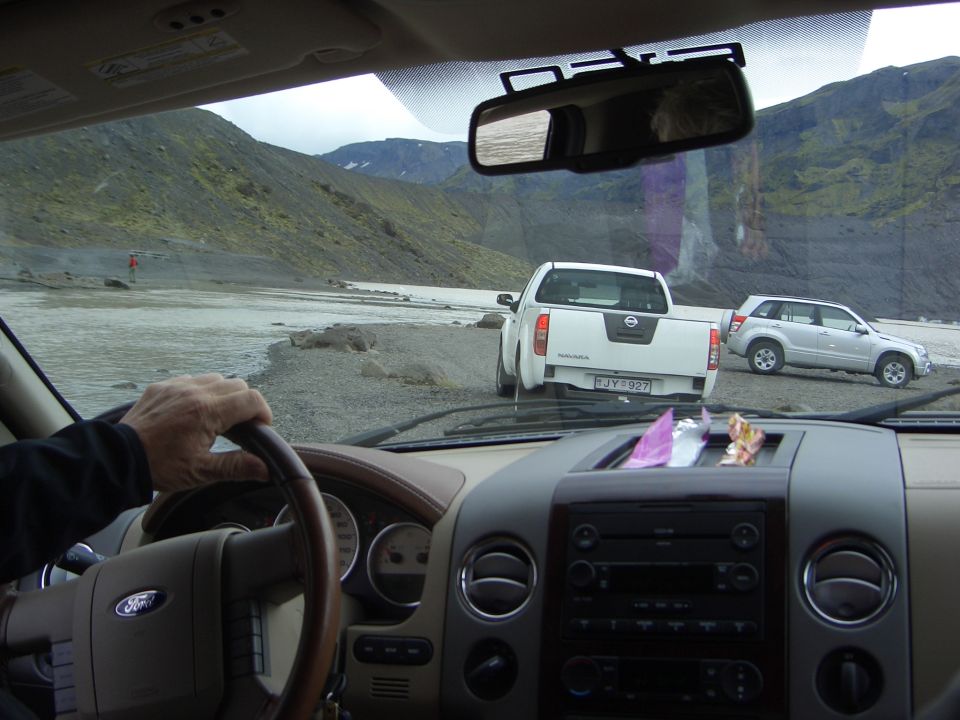 View from inside a vehicle shows a white Nissan pickup and a silver SUV parked on a gravel shore near a river, with mountains behind