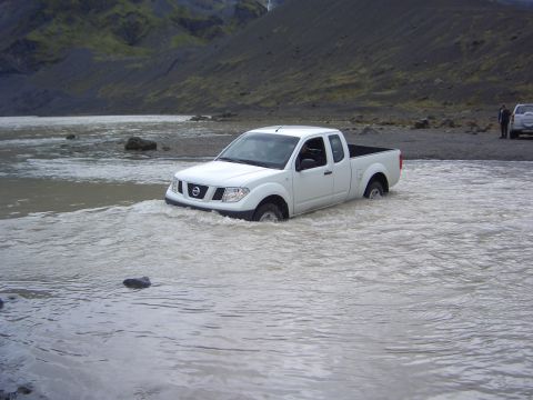 1974 Ford Bronco - A white pickup truck navigates through a shallow body of water, with a landscape in the background.