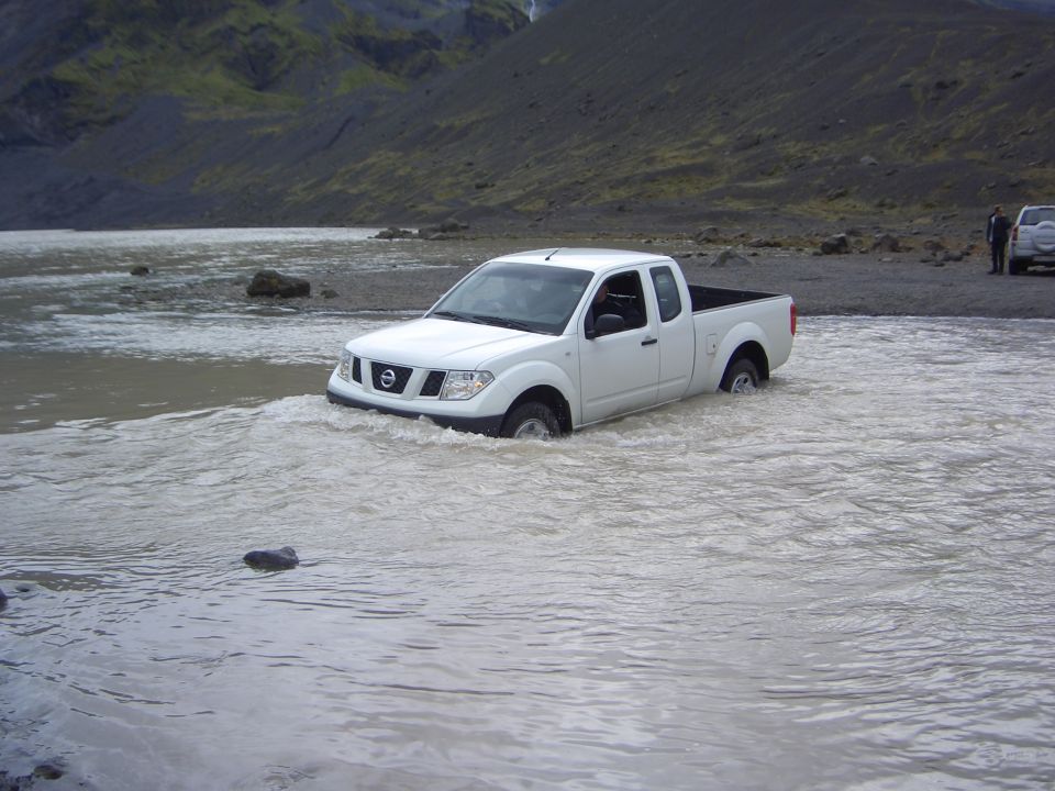 White Nissan pickup truck driving through shallow water with a rocky, mountainous background. Another white vehicle is parked on the shore