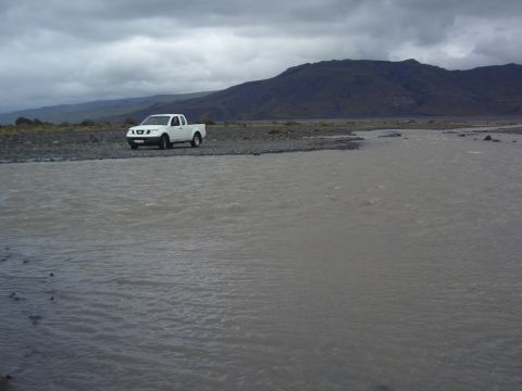 1974 Ford Bronco - A white pickup truck is parked on the bank of a wide, shallow river with mountains in the background.
