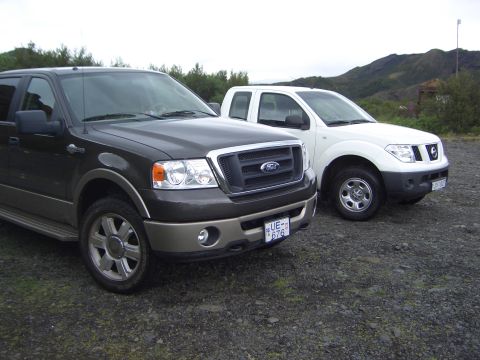 1974 Ford Bronco - A dark Ford F-150 and a white Nissan pickup are parked side-by-side on a gravel lot with a mountainous background.