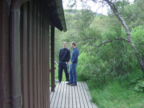 1974 Ford Bronco - Two men converse on a wooden deck outside a rustic structure, surrounded by dense greenery and trees.