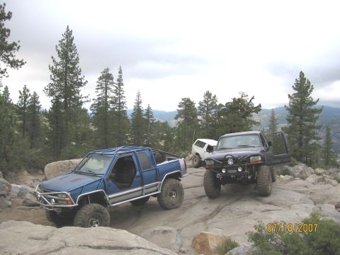 1994 Ford Bronco - Three off-road trucks navigate a challenging rocky terrain under a cloudy sky.