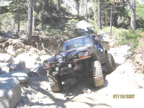 1994 Ford Bronco - A modified truck is shown driving on a rough, rocky path through a wooded area.