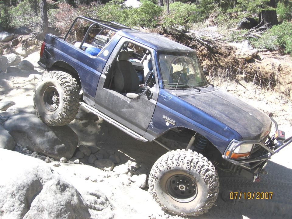 Doorless blue Ford Bronco with roll cage crawling over granite boulders on a trail