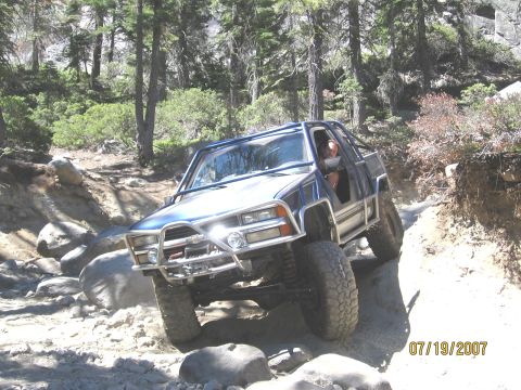 1994 Ford Bronco - A modified pickup truck navigates a challenging off-road path through a wooded area.