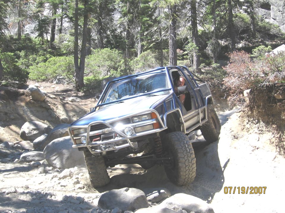 Caged blue 4-door SUV with winch bumper flexing over boulders on a pine forest rock trail