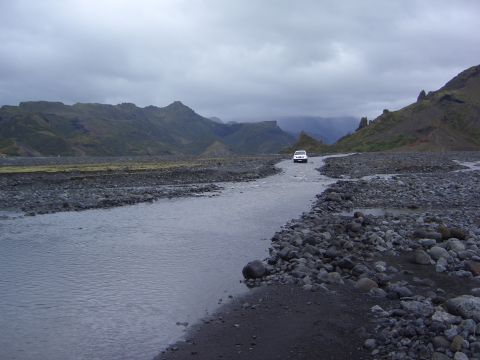 1974 Ford Bronco - A lone white SUV navigates a rocky riverbed amidst a dramatic, overcast mountain vista.