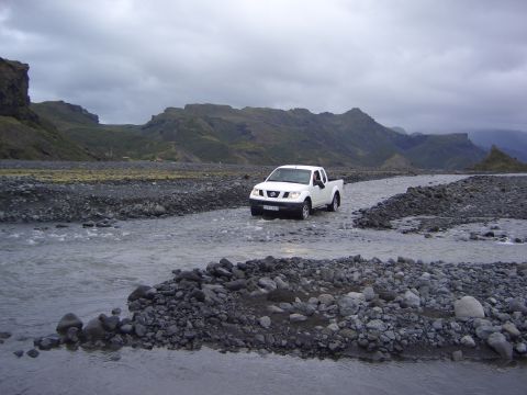 1974 Ford Bronco - A white pickup truck navigates a shallow river in a , mountainous landscape under an overcast sky.