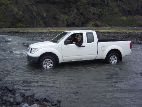 1974 Ford Bronco - White pickup truck navigates a shallow river, with a person visible in the driver's seat.