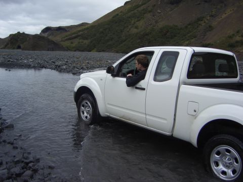 1974 Ford Bronco - A white pickup truck navigates a rocky stream bed under a cloudy sky.