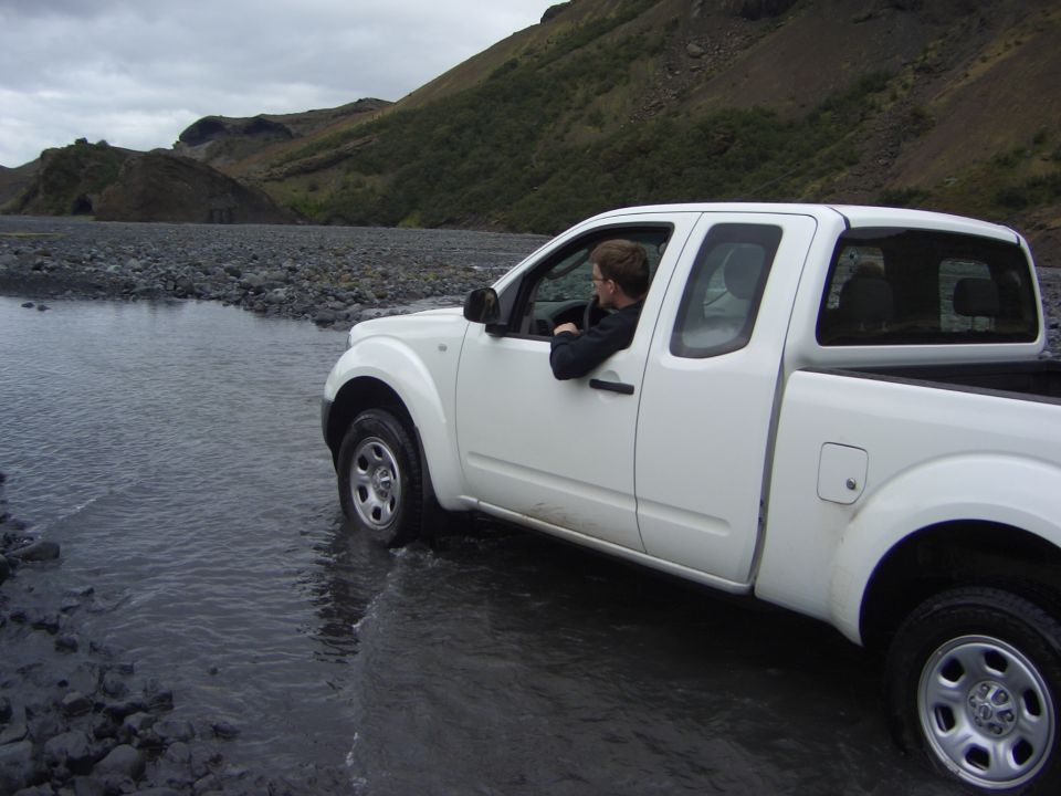 Person driving a white pickup truck through a shallow, rocky stream with hills in the background