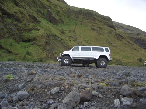 1974 Ford Bronco - A modified white van with large off-road tires sits on a rocky terrain with a green, mossy hill in the background.