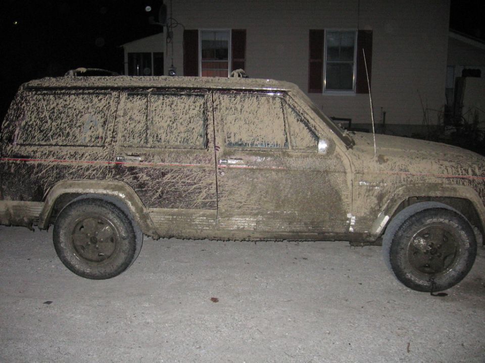 Mud-covered 1993 Jeep Cherokee, side view. Dark red paint visible under mud. Antenna extended