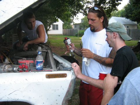 1990 Ford Bronco - Three men appear to be working on the engine of a white Ford Bronco, with tools and a beer bottle visible.
