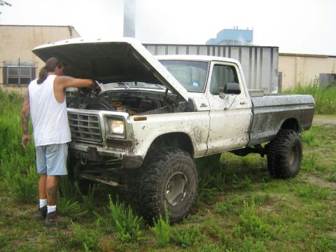 1990 Ford Bronco - A man inspects the engine of a lifted, mud-covered pickup truck parked in a grassy area.