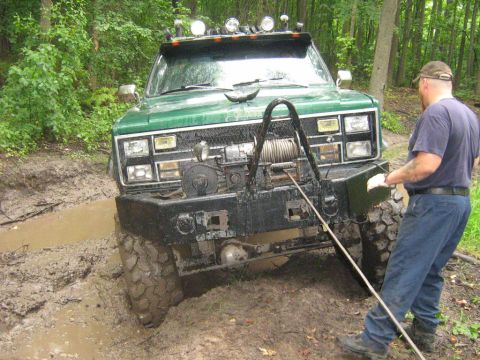 1990 Ford Bronco - A man attempts to free a green off-road truck that is stuck deep in the mud on a wooded trail.