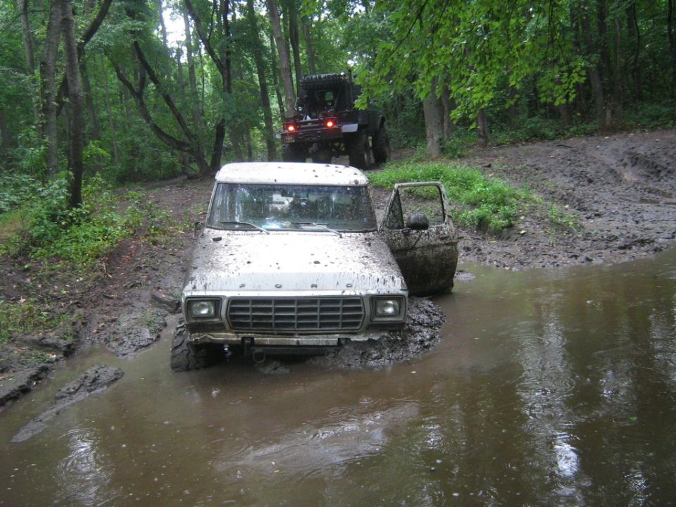 Mud-splattered white Ford Bronco with open driver's door stuck in a muddy puddle, another off-road vehicle behind it