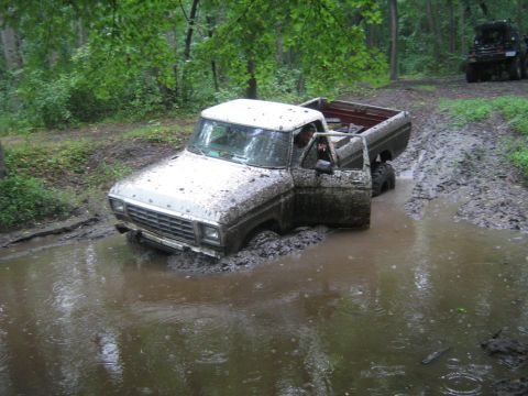1990 Ford Bronco - A white Ford F-Series truck is stuck in a muddy puddle off-road, with another vehicle visible in the background.