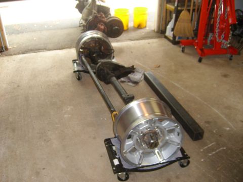 1994 Ford Bronco - A front axle assembly with two large alloy wheels is positioned on dollies in a garage setting, with tools and equipment visible in the bac…