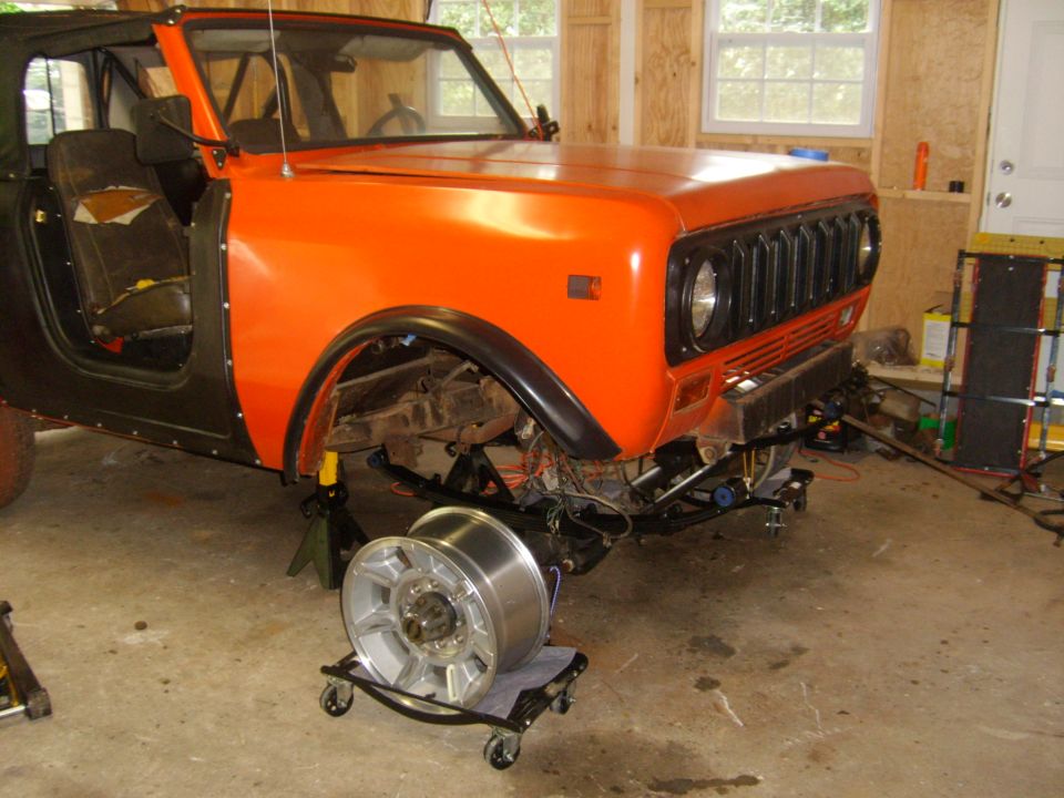 Orange International Harvester Scout II with black fenders, missing front wheel, on jack stands in a garage
