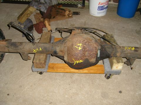1994 Ford Bronco - A rusty rear axle assembly is shown, supported by wooden blocks on a wheeled dolly. Yellow markings are visible on the housing.
