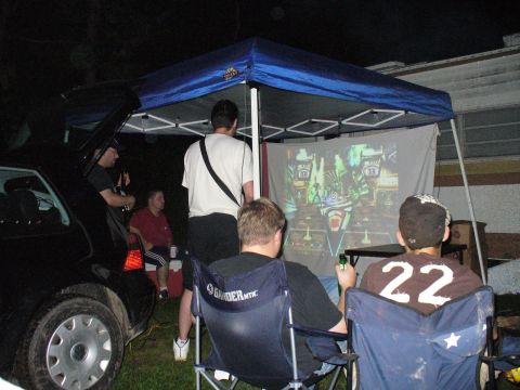 1991 Ford Probe - People gather outdoors at night to watch a projected image, with a black sedan parked nearby.