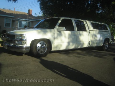 1990 Ford Bronco - white custom stretched Chevrolet dually pickup with dual rear wheels and a camper shell, parked in a residential driveway.