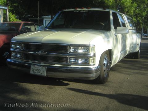 1990 Ford Bronco - white Chevrolet dually truck with a billet grille, chrome bumper and lowered stance, parked near a red car.