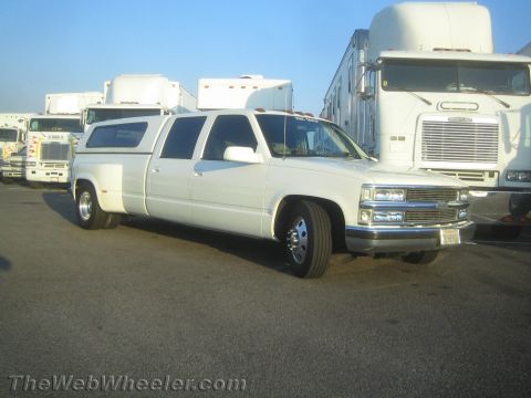 1990 Ford Bronco - white custom stretched Chevrolet dually truck with a camper shell and dual rear wheels, parked in a lot among semi trucks.