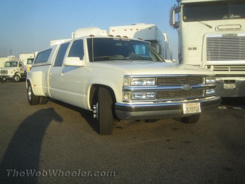 1990 Ford Bronco - white custom stretched Chevrolet dually truck with billet grille and dual rear wheels, parked in a yard beside semi trucks.