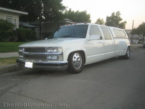1990 Ford Bronco - white custom stretched Chevrolet dually truck with a camper shell and dual rear wheels, parked on a residential street at dusk.