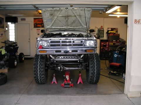 1986 Toyota Hilux - Head-on view of a white Toyota pickup in a garage with the hood up, showing a chrome grille and bumper, a newly fitted solid front axle wit…