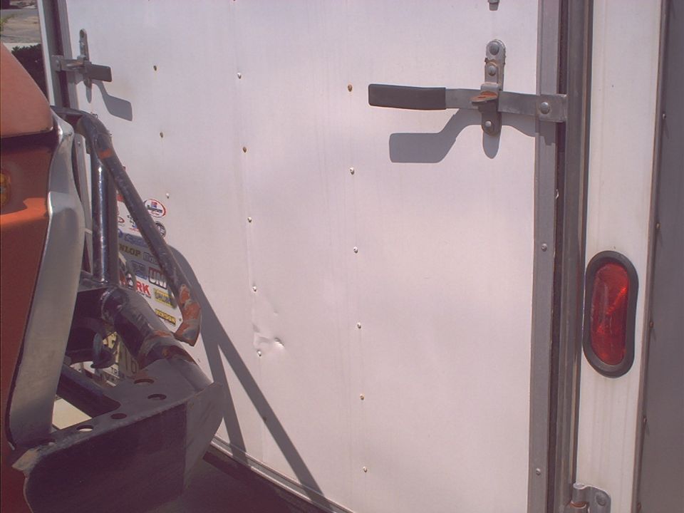 Trailer door latch mechanism on a white trailer, with part of an orange object and black metal frame visible