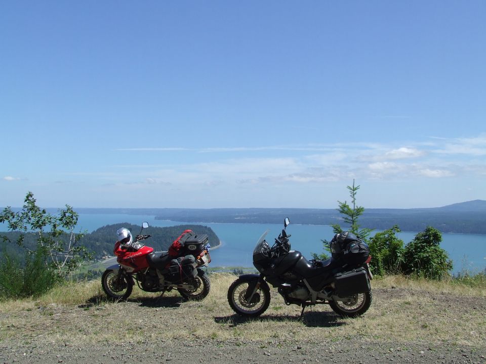 Two touring motorcycles parked on a grassy overlook with a bay and distant hills in the background