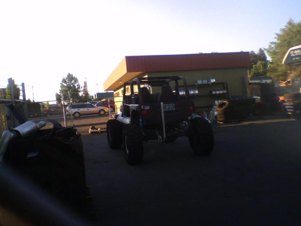 Modified off-road vehicle with large tires, rear view, dark color, parked near a building