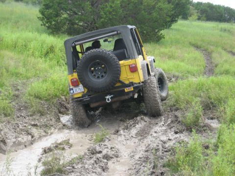 1986 Ford Bronco - Yellow Jeep Wrangler navigates a challenging muddy off-road path.