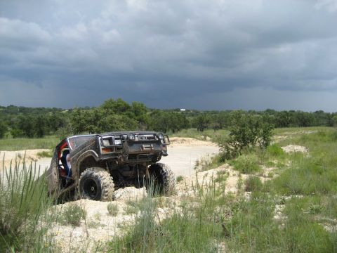 1986 Ford Bronco - An off-road vehicle navigates a challenging dirt track, its tires caked in mud, beneath a dramatic, cloud-filled sky.