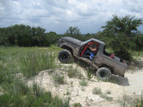 1986 Ford Bronco - This modified off-road vehicle navigates a challenging incline on a dirt trail under a cloudy sky.