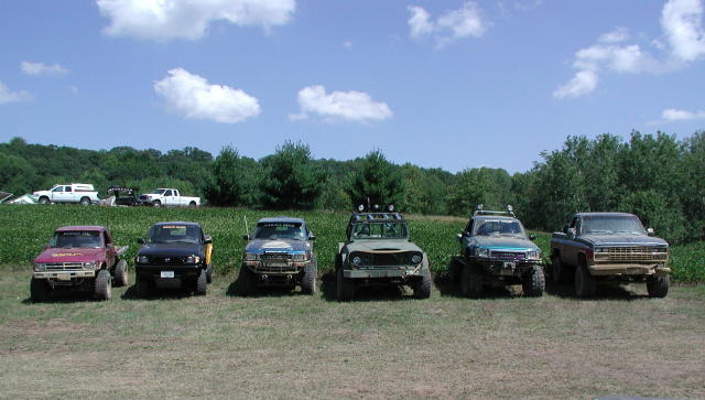 Row of six lifted 4x4 mud trucks parked front-facing in a field by a cornfield