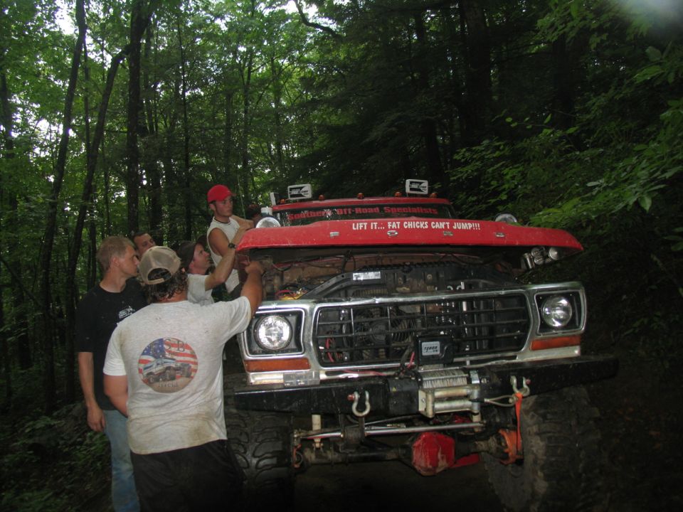 Several guys looking at the engine of a red off-road truck in a wooded area. The hood is up