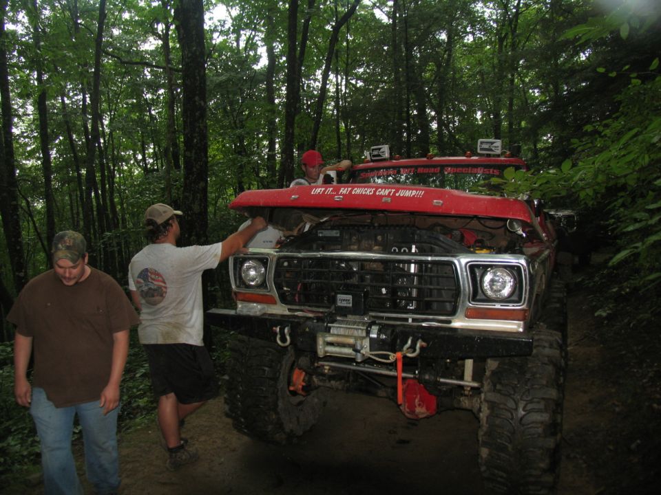 Red off-road truck with hood open, winch, and off-road tires. Three men are visible