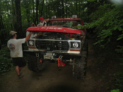 1995 Ford C-350 (Centurion Conversions) - Off-road enthusiasts work on a red truck in a wooded area, with a humorous sticker on the hood.