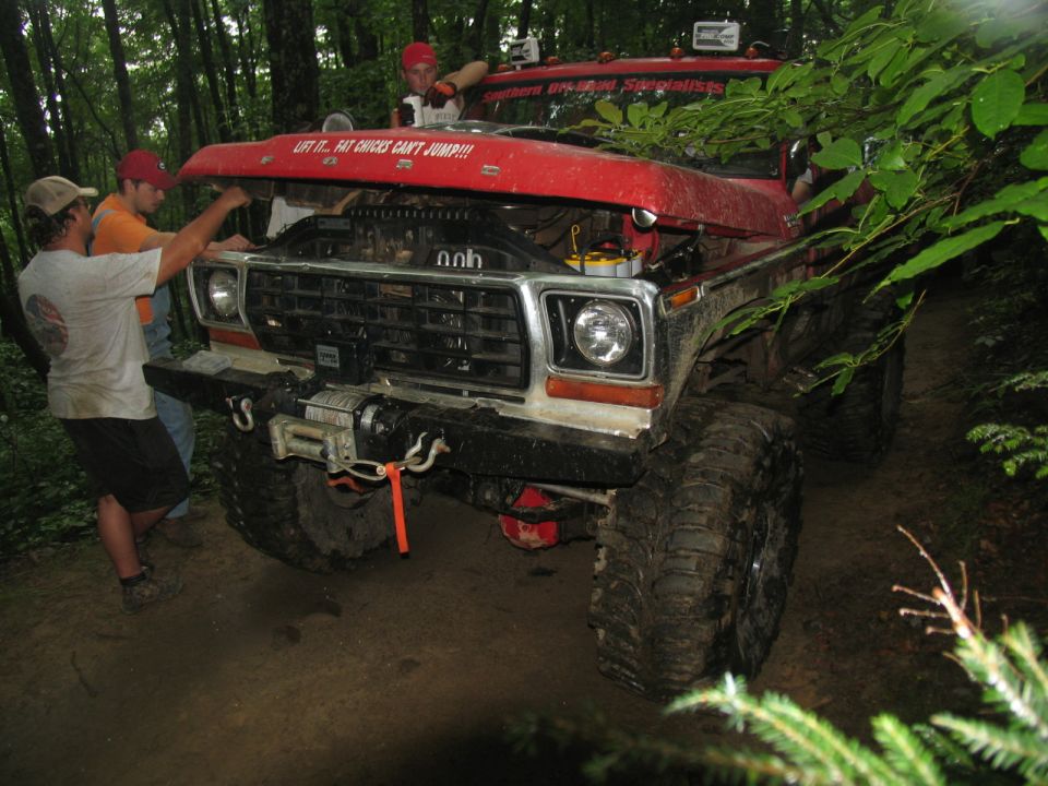 Red off-road truck with hood open, winch on front bumper. Three people are looking at the engine