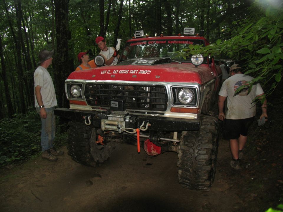 Red Ford F-Series truck with large tires and winch, people around it in woods