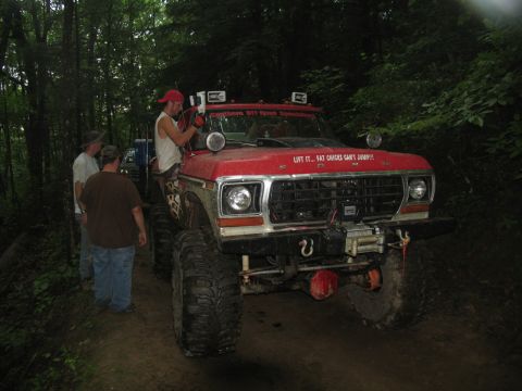 1995 Ford C-350 (Centurion Conversions) - A modified red truck with oversized tires and a winch is parked on a dirt road surrounded by trees.