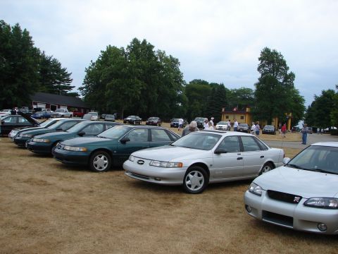 1993 Ford Taurus - A gathering of sedans parked on a grassy field under a cloudy sky.