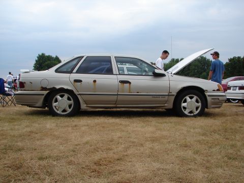 1993 Ford Taurus - A tan 1993 Ford Taurus with significant rust damage on its lower body panels is displayed outdoors.
