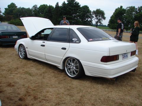 1993 Ford Taurus - White 1993 Ford Taurus at a car show, hood up, with custom wheels and decals on the rear window.