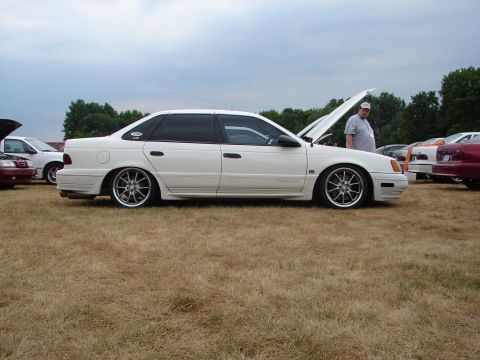 1993 Ford Taurus - A white sedan sits on a grassy field with its hood open, surrounded by other vehicles.